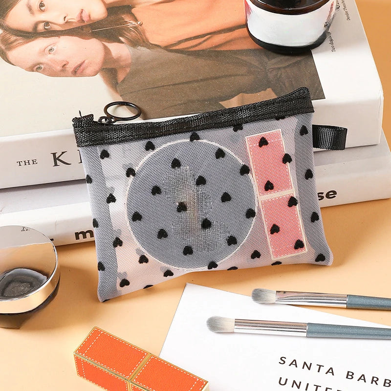A clear mesh cosmetic storage pouch with black and pink accents resting on a wooden surface next to makeup brushes.