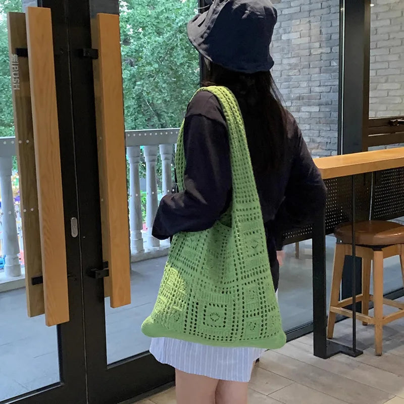 A woman wearing a dark top and light skirt holds a bright green knitted shoulder bag in front of a glass door.