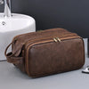 A brown PU leather travel toiletry bag with organizer pockets resting on a gray surface next to a white sink.
