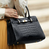 A black genuine leather shoulder bag with gold hardware carried by a person wearing a beige top and skirt.