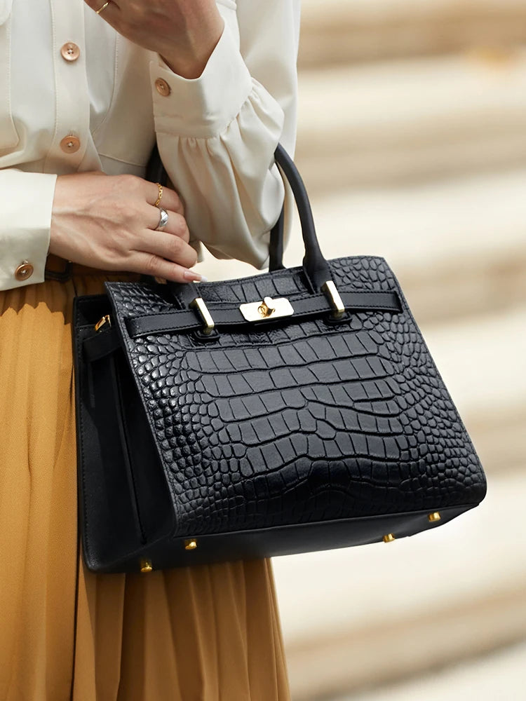 A black genuine leather shoulder bag with gold hardware carried by a person wearing a beige top and skirt.