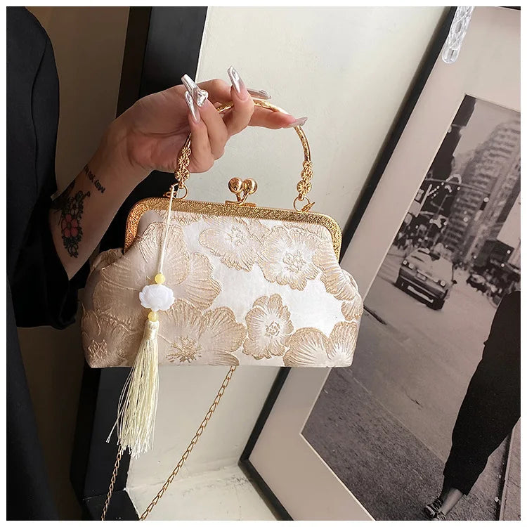 A hand holds an embroidered silk evening clutch with a gold chain strap against a light background.