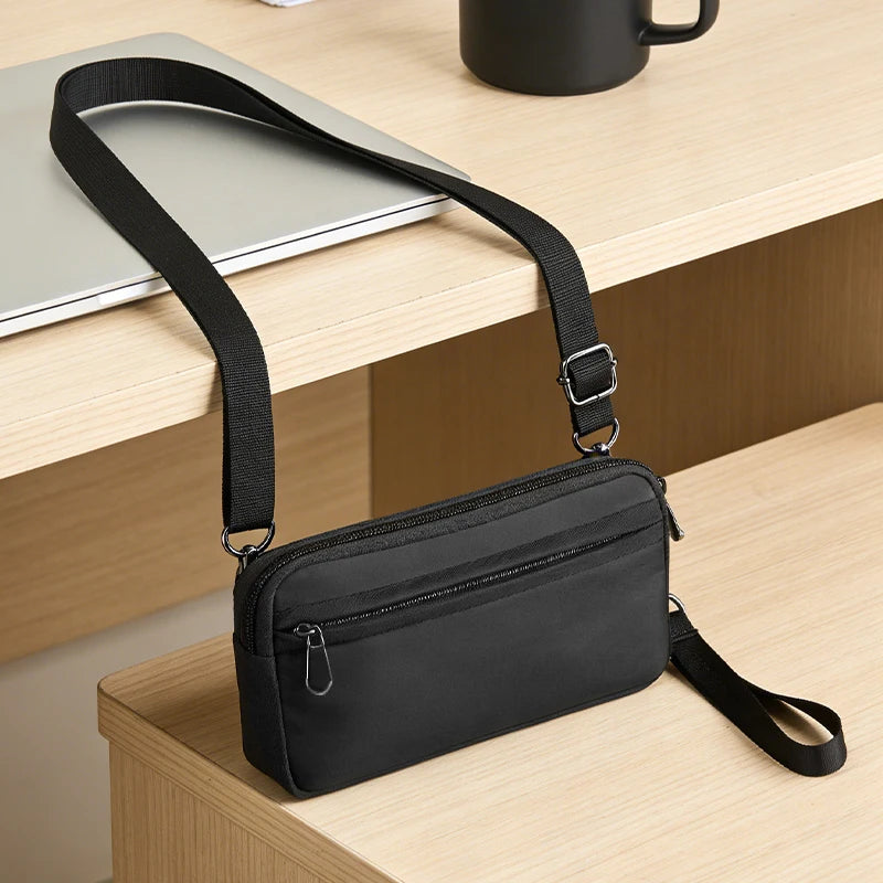 A black zippered phone pouch with a strap is resting on a light wood surface next to a laptop.