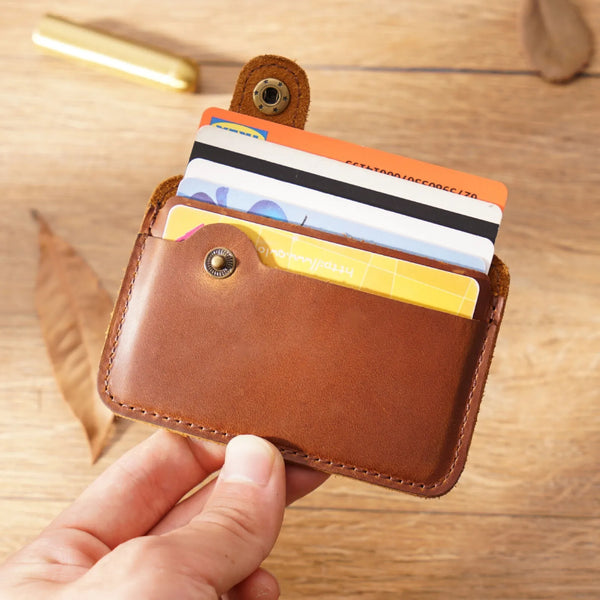 A hand holds a brown genuine leather slim card holder containing several credit cards on a wooden surface.