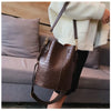 A brown alligator pattern leather crossbody bag is displayed hanging on a person next to a gray sofa.