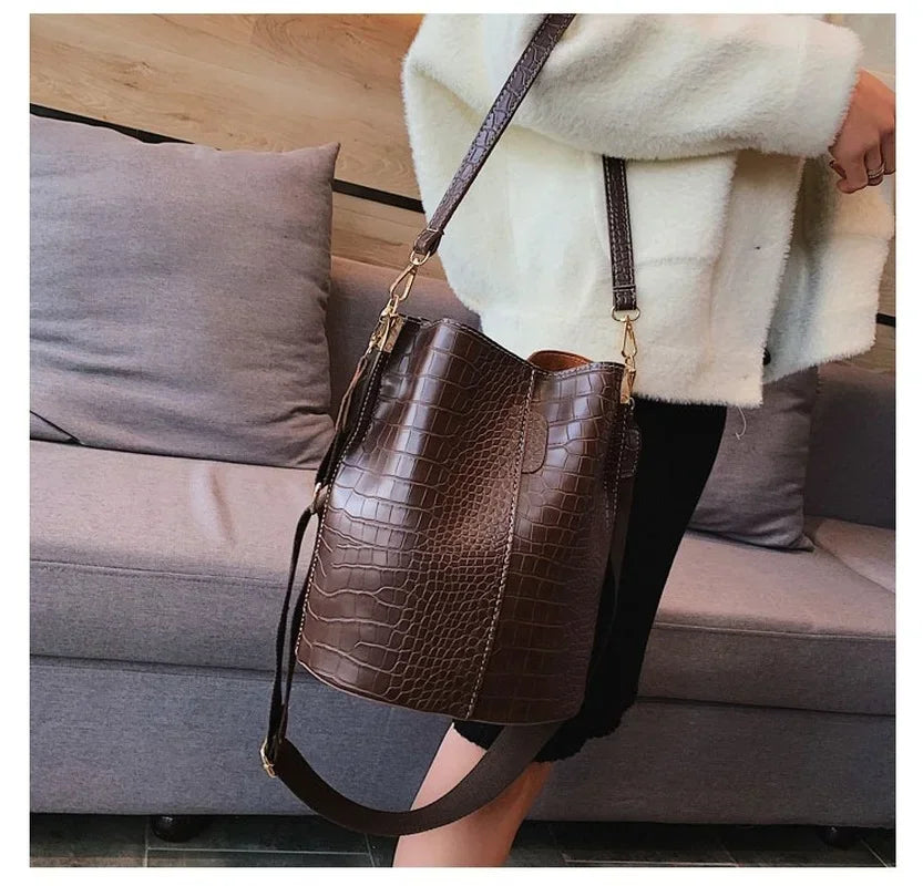 A brown alligator pattern leather crossbody bag is displayed hanging on a person next to a gray sofa.
