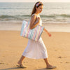 A woman walks on a beach holding a striped transparent PVC tote bag.