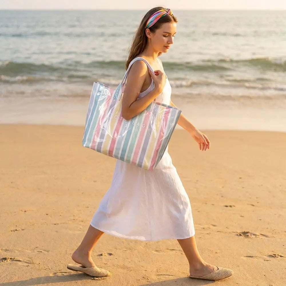A woman walks on a beach holding a striped transparent PVC tote bag.