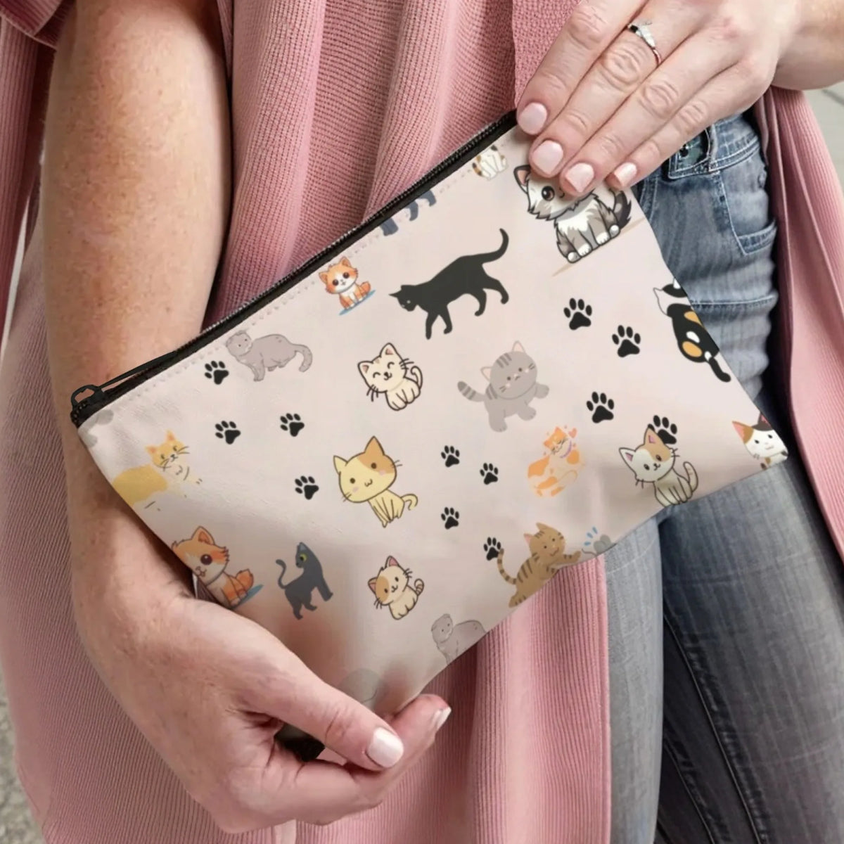 A hand holds a beige cartoon cat cosmetic pouch with various cat illustrations against a pink cardigan.