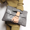 A hand holding a gray leather crossbody clutch bag against a beige sweater and olive green pants.