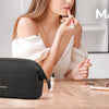 A woman holds a black padded makeup bag on a wooden table with various beauty products nearby.