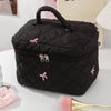 A black quilted cosmetic bag with a handle and zipper, featuring pink bow accents, resting on a white surface.