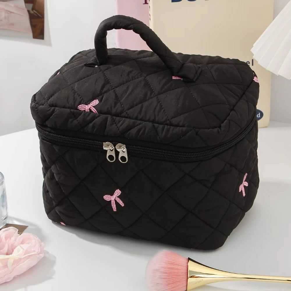 A black quilted cosmetic bag with a handle and zipper, featuring pink bow accents, resting on a white surface.