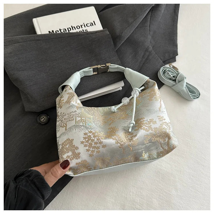 A light gold embroidered fabric crossbody bag resting on a dark gray blazer with a white notebook.
