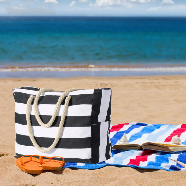 Un sac de plage rayé noir et blanc posé sur le sable avec un livre ouvert et une serviette bleue.