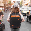 A person wearing a hat is carrying a black expandable travel backpack with orange accents on a city street.