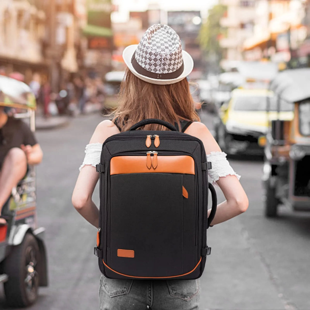 A person wearing a hat is carrying a black expandable travel backpack with orange accents on a city street.