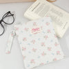 A white floral plaid cotton tablet pouch with a pink tag and a light-colored notebook nearby.