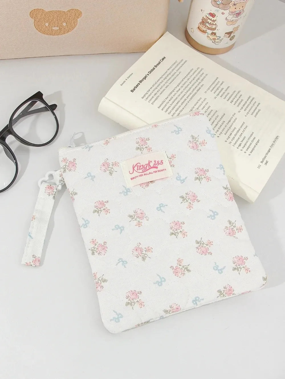 A white floral plaid cotton tablet pouch with a pink tag and a light-colored notebook nearby.