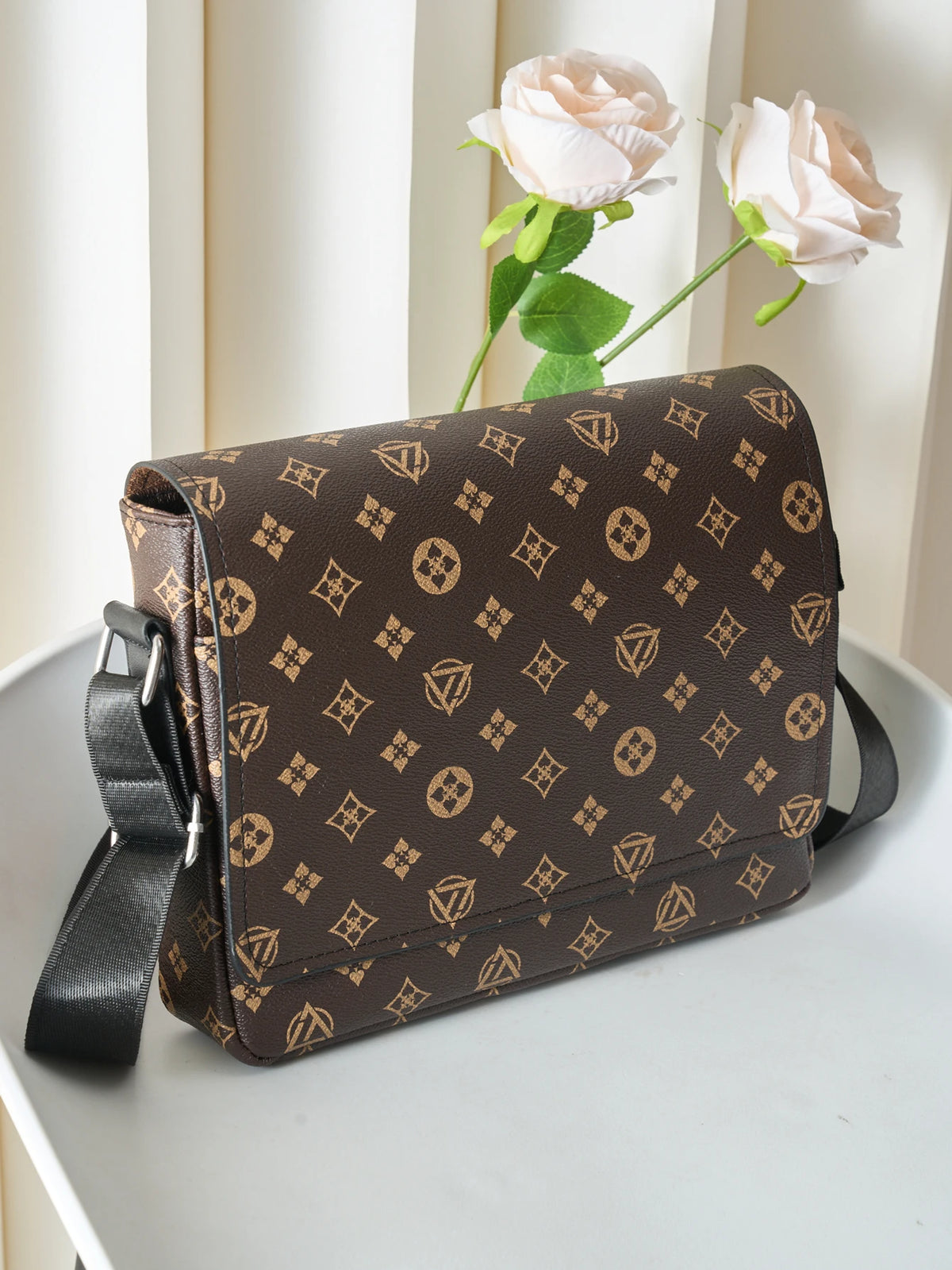 A brown men's casual crossbody bag with a patterned fabric rests on a white surface next to a pink rose.