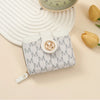 A light gray patterned compact wallet with a leather strap rests on a beige surface next to a watch and a dried flower.