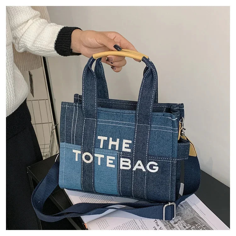 A dark blue denim tote bag with white stitching and a light blue accent, held by a hand.