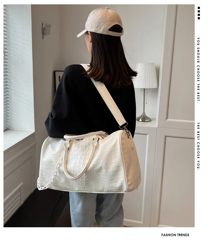 A woman wearing a black top and light pants holds a cream and beige crocodile grain PU leather travel duffle bag.