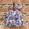 A purple and white reusable shopping bag featuring cute animal graphics against a brick background.