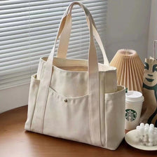 A beige canvas tote bag with light tan straps resting on a wooden surface next to a potted plant.