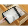 A hand holds a large transparent woven storage bag with black straps inside a wooden drawer.