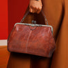 A brown leather crossbody bag with silver hardware is held by a person against a red background.