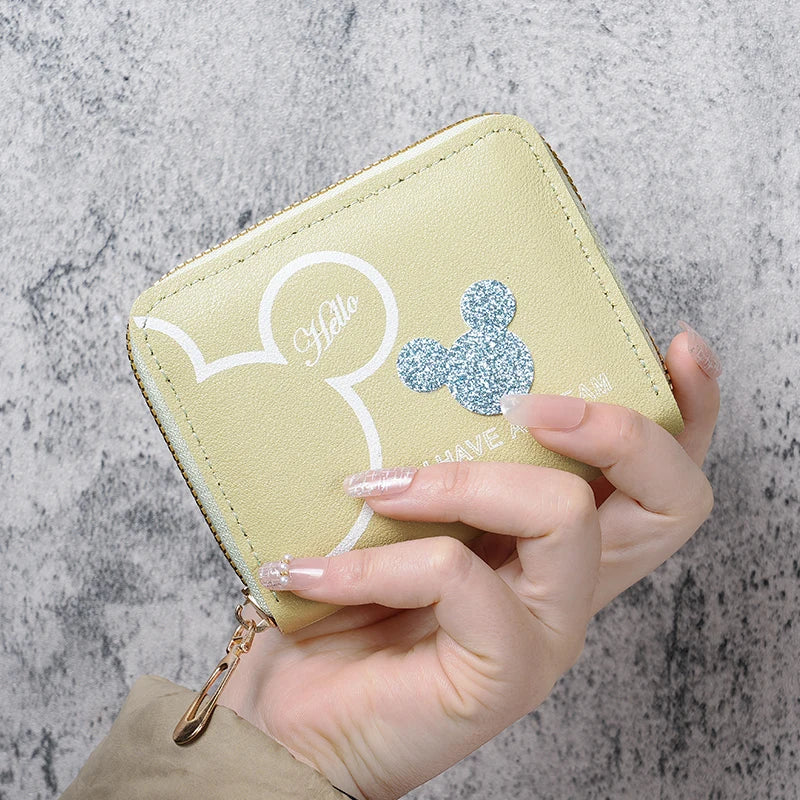 A hand holds a light green cartoon Mickey Mouse zipper coin purse against a gray textured background.
