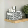 A gray and white patterned foldable cotton linen storage basket with handles sits on a wooden shelf next to a potted green pl