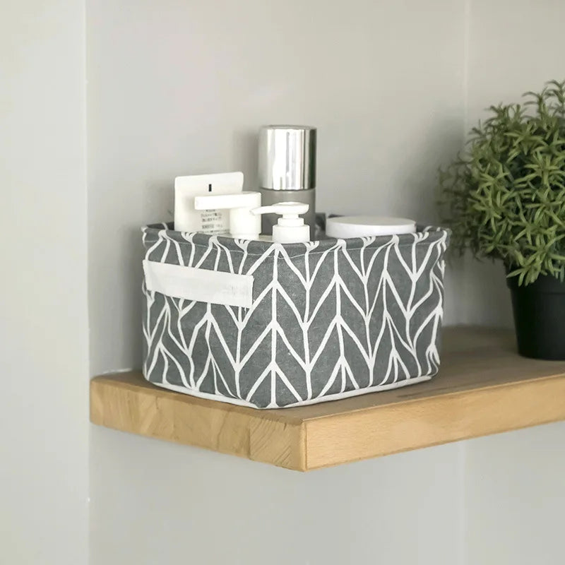 A gray and white patterned foldable cotton linen storage basket with handles sits on a wooden shelf next to a potted green pl