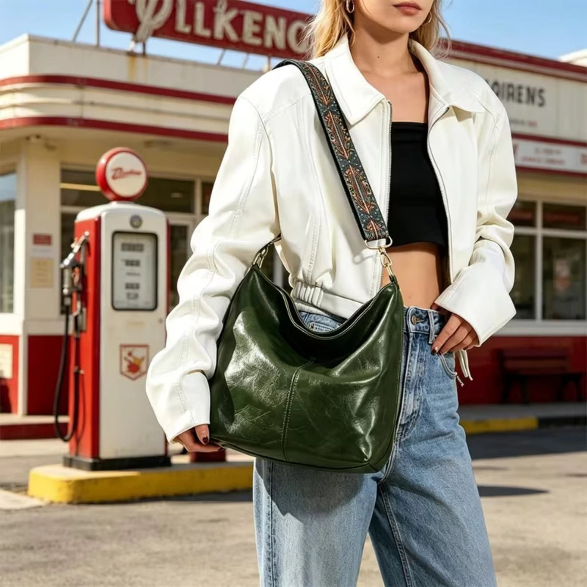 A woman wearing a white jacket and jeans holds a green leather crossbody bag with a patterned strap.
