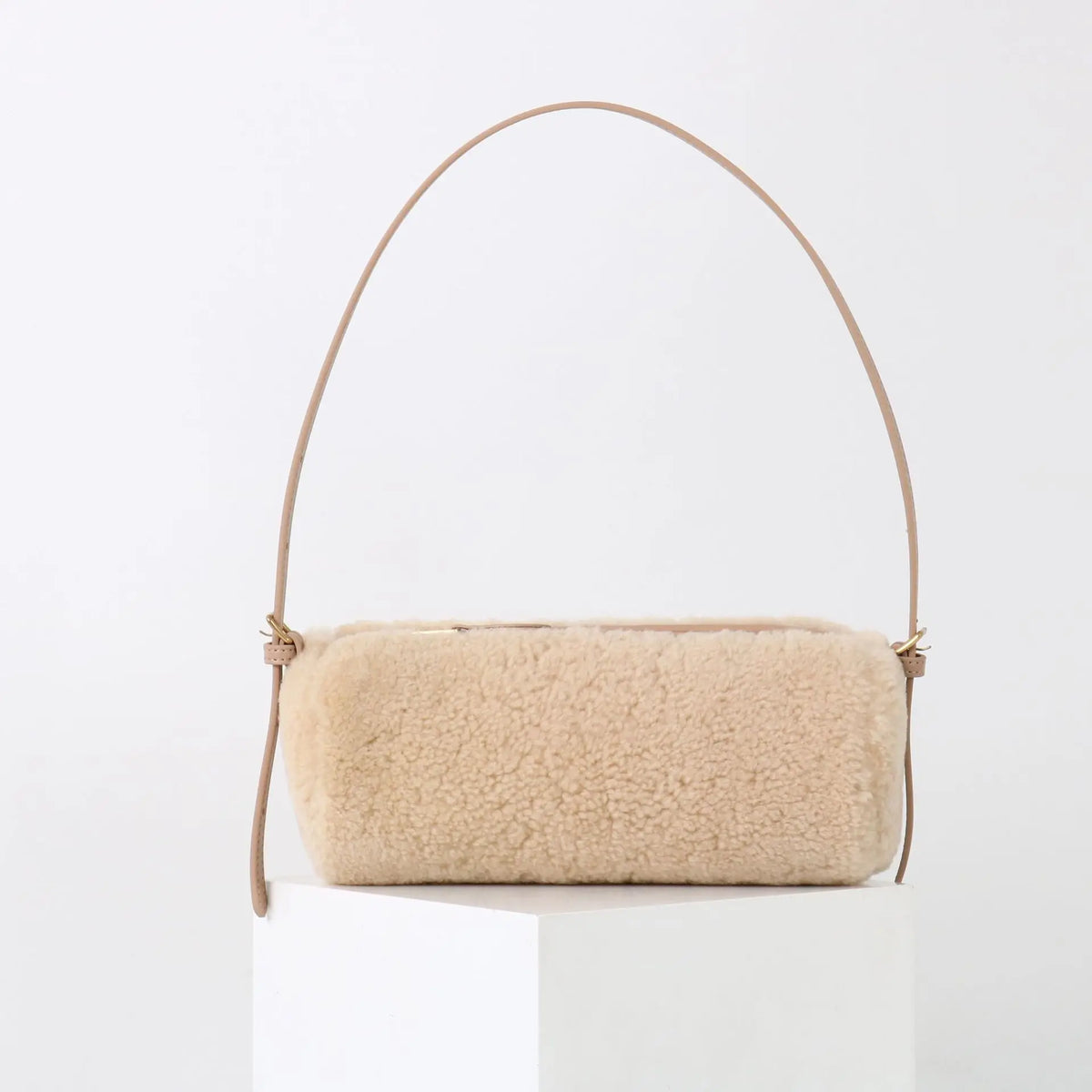 A beige vintage cow leather cylinder shoulder bag with a thin tan strap displayed on a white pedestal.
