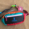Bunte Outdoor-Sport-Crossbodytasche mit orange, pink und grün Akzenten auf tanfarbiger Stoffoberfläche.