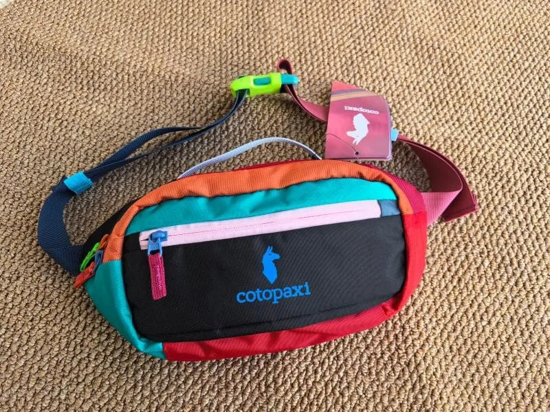 Bunte Outdoor-Sport-Crossbodytasche mit orange, pink und grün Akzenten auf tanfarbiger Stoffoberfläche.
