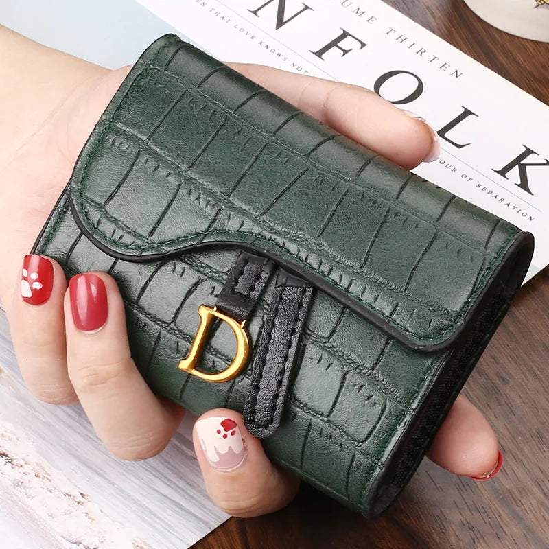 Une main tient un porte-cartes clutch vert foncé texturé avec une initiale dorée.