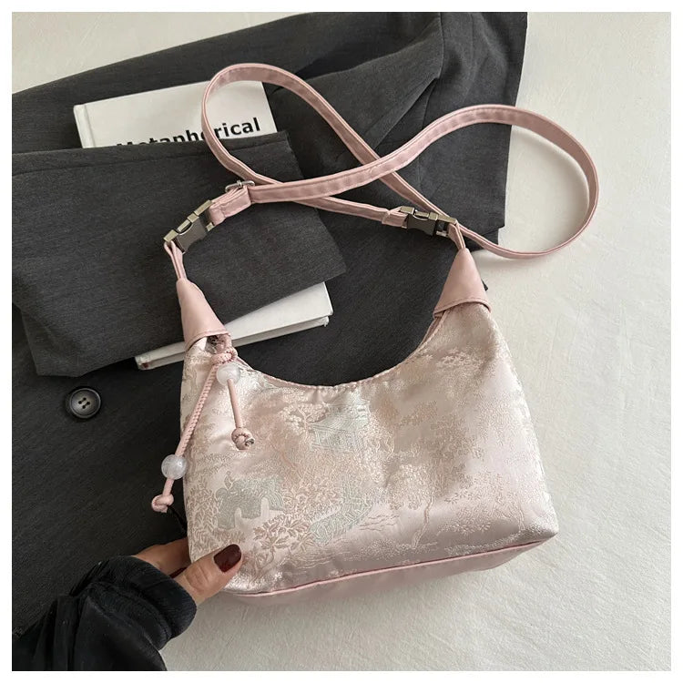 A light pink satin embroidered crossbody shoulder bag is displayed against a dark gray blazer and white book.