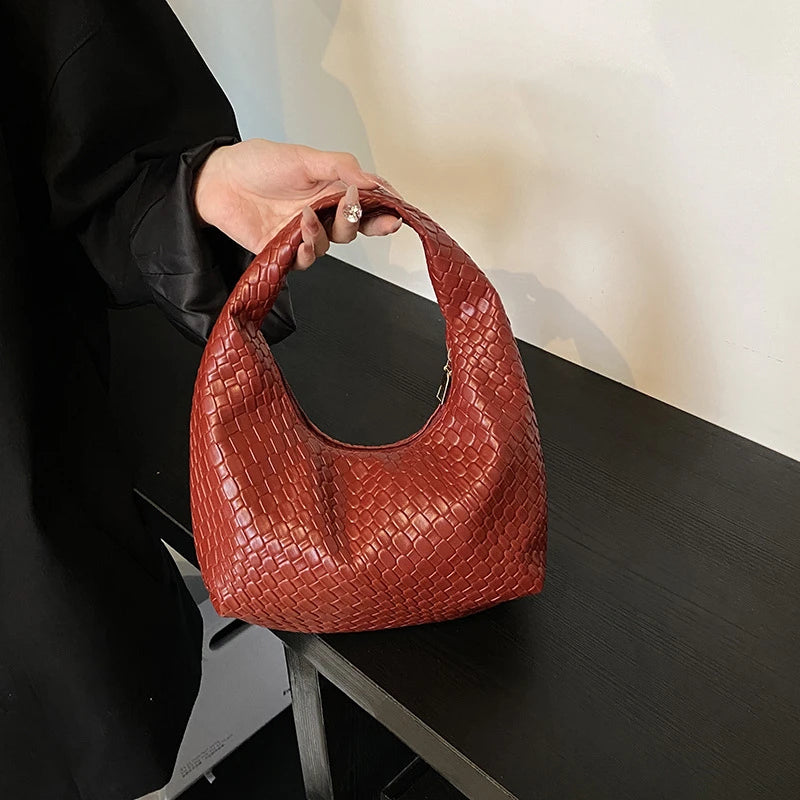 A close-up of a reddish-brown woven embossed hobo shoulder bag with a top handle resting on a dark table.