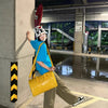 A person wearing a blue shirt and khaki pants is holding a yellow faux crocodile tote bag in an indoor public area.