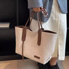 A beige canvas shoulder tote bag with brown leather handles is held by a person wearing a light jacket and white pants.
