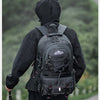 A person wearing a black waterproof hiking backpack is shown from the back, standing on a path with greenery in the backgroun