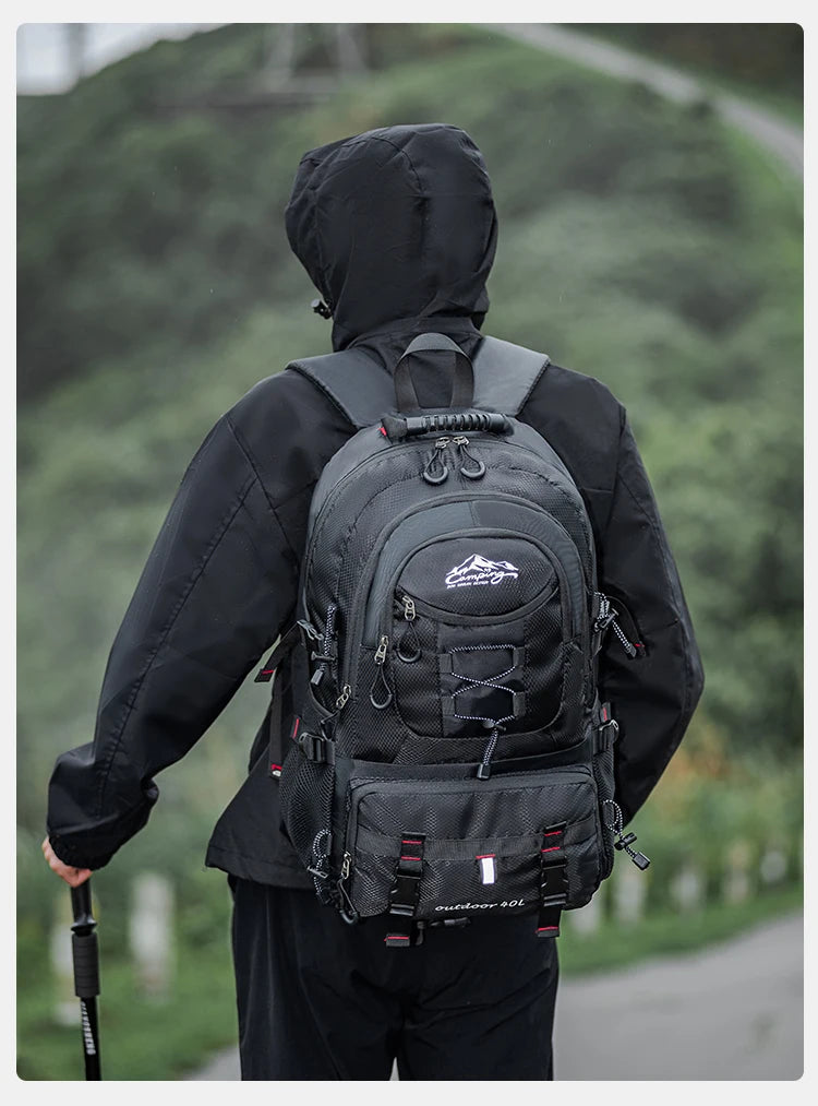 A person wearing a black waterproof hiking backpack is shown from the back, standing on a path with greenery in the backgroun