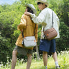 A young man and woman walk through a green outdoor area, with the man carrying a brown crossbody bag.