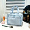 A light blue and white checkered multipocket travel toiletry bag with fabric handles sits on a white surface.