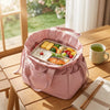 A pink insulated drawstring lunch bag filled with various food items on a wooden table.