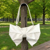 A white pleated bow crossbody tote bag is displayed hanging on a tree trunk in a park setting.