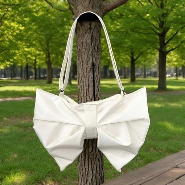 A white pleated bow crossbody tote bag is displayed hanging on a tree trunk in a park setting.